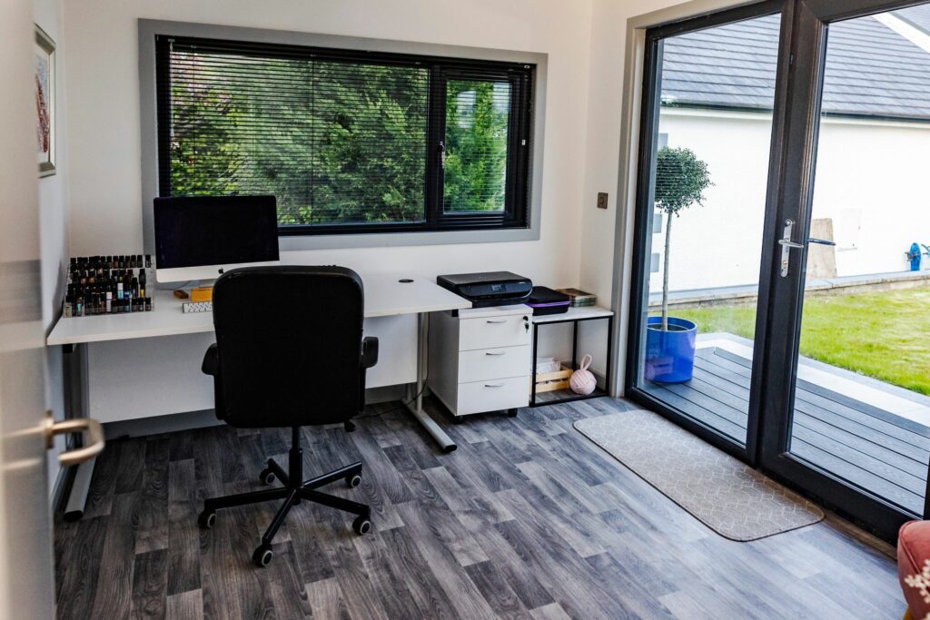 Modern home office with desk, computer, and chair, overlooking a garden through large glass doors.