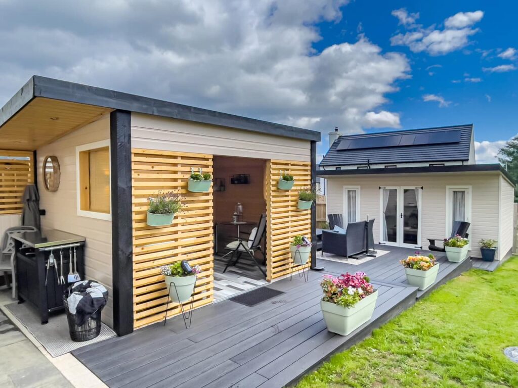 Modern patio and small house with solar panels, surrounded by planters and outdoor seating, under a partly cloudy sky.