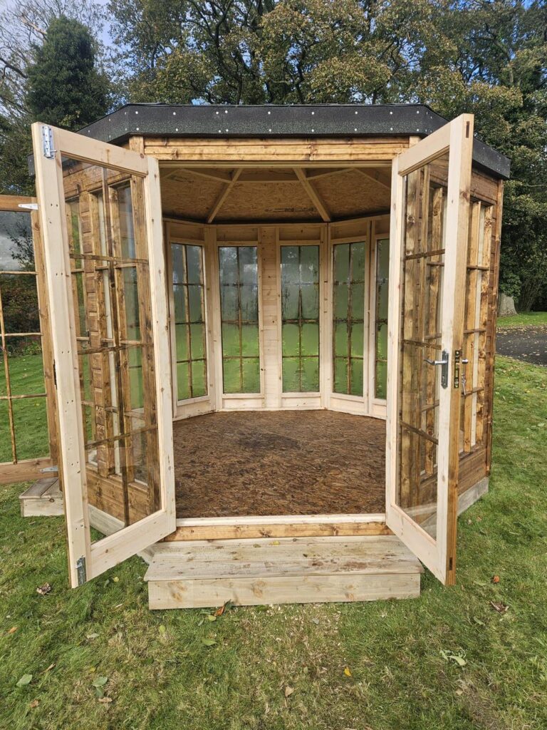 Wooden garden gazebo with open double glass doors, set in a grassy area.