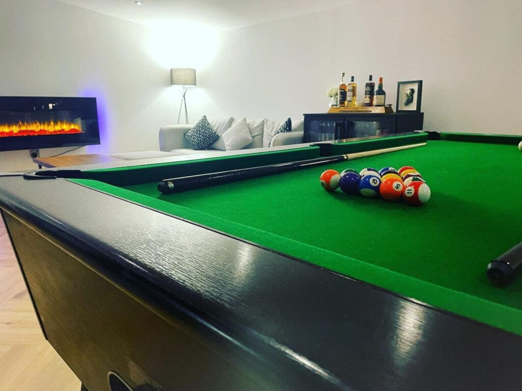 Pool table in a cozy living room with green cloth, cue sticks, and billiard balls, next to a modern electric fireplace and seating area.