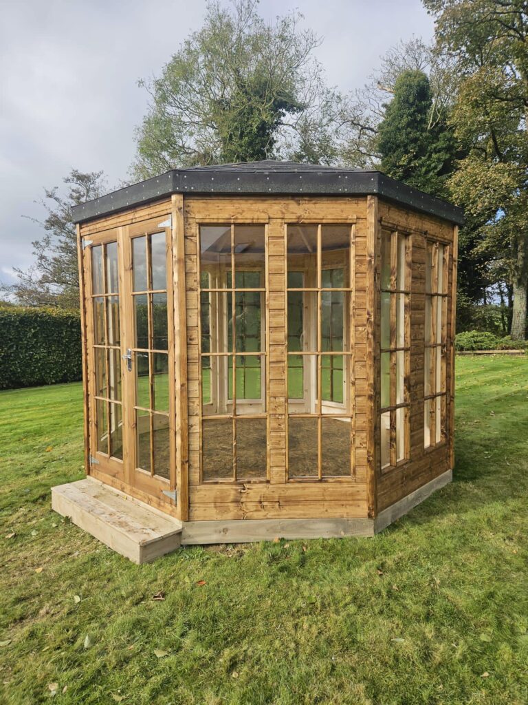Wooden garden gazebo with large windows on a grassy lawn, surrounded by trees.