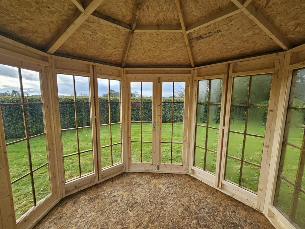 Interior of a wooden gazebo with large windows overlooking a grassy garden.