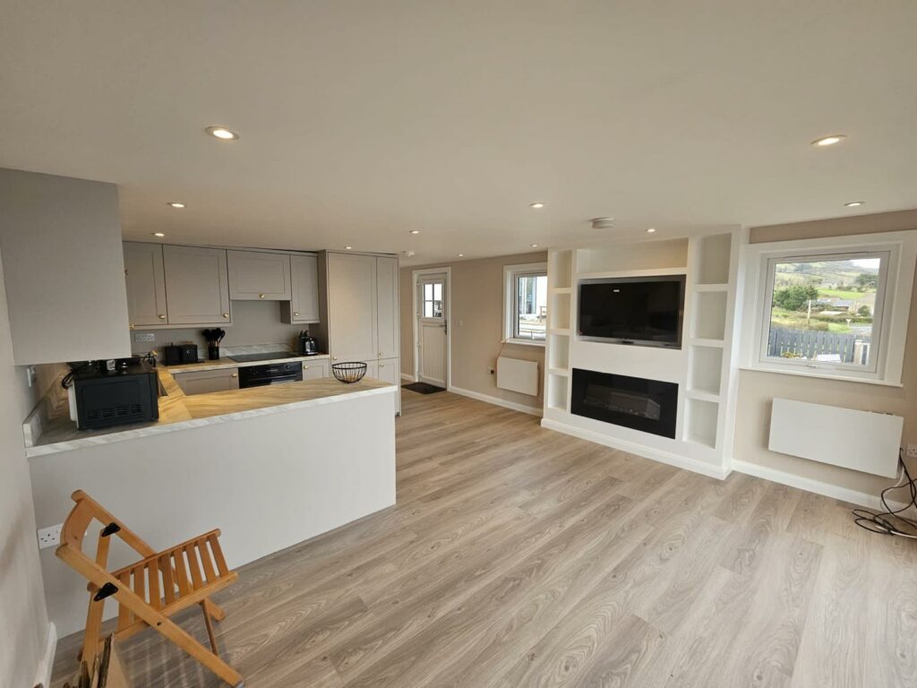Modern studio apartment with open-plan kitchen, wood flooring, built-in TV and fireplace, and large windows.