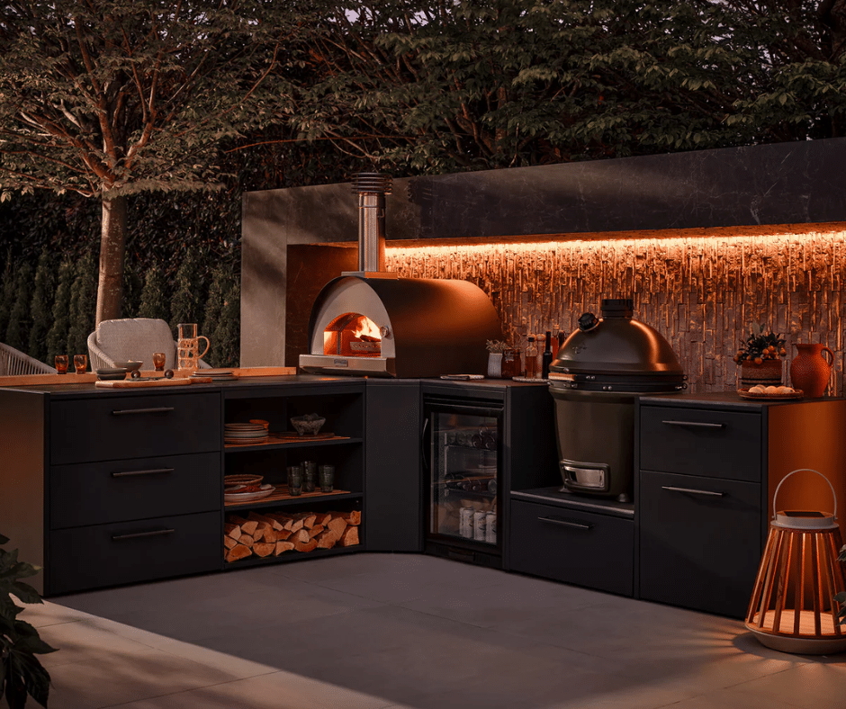 Outdoor kitchen with pizza oven, grill, and fridge, surrounded by modern cabinetry and subtle lighting.
