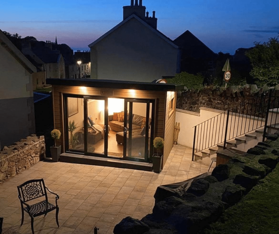 Cozy lighted cabin with large windows, patio seating, and garden view at dusk.