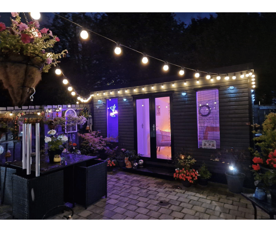 Cozy patio with string lights, flowers, and a small cabin illuminated in purple, creating a tropical ambiance at night.