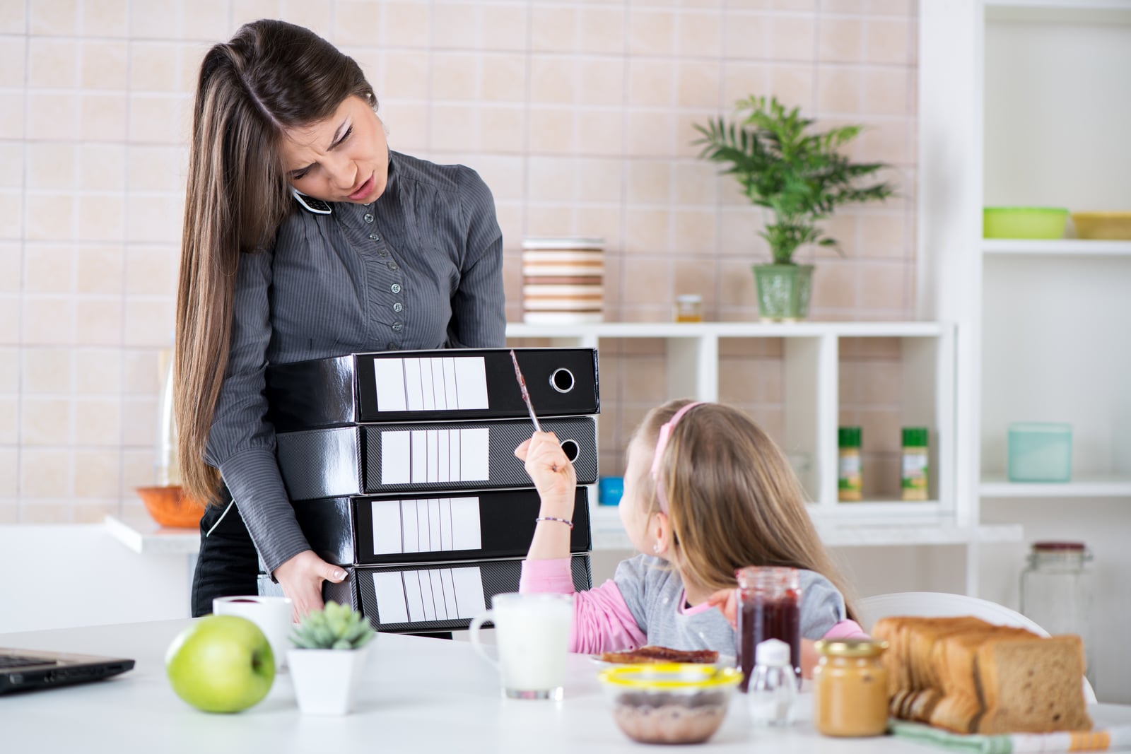 Mother multitasking with folders and phone while daughter points at breakfast table.